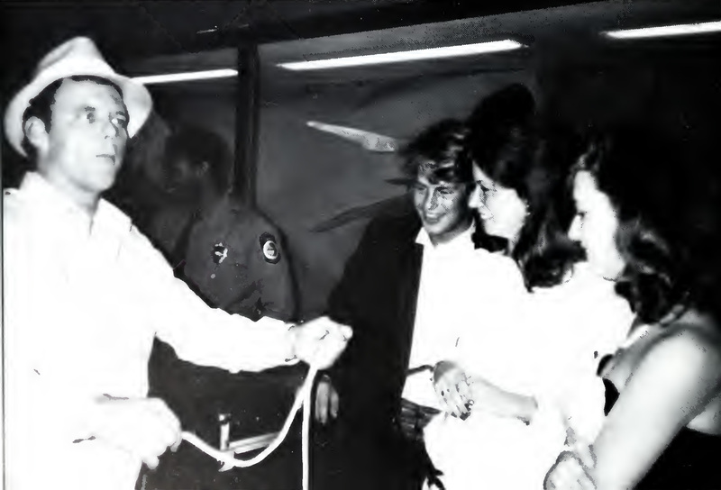 This is a photo of three students in black tie attire at the 1987 Fancy Dress dance with a man who is showing them a rope.