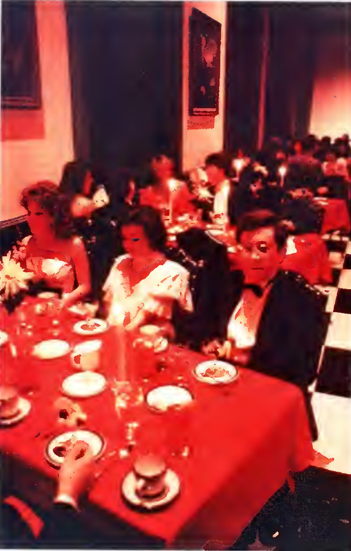 This is a photograph of the dinner held during the 1985 Fancy Dress at W&L. There is theme-specific decor, including paintings and red tablecloths. At the table, there are several white plates and guests sitting at it.