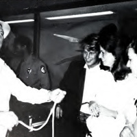 This is a photo of three students in black tie attire at the 1987 Fancy Dress dance with a man who is showing them a rope.