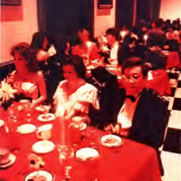 This is a photograph of the dinner held during the 1985 Fancy Dress at W&L. There is theme-specific decor, including paintings and red tablecloths. At the table, there are several white plates and guests sitting at it.